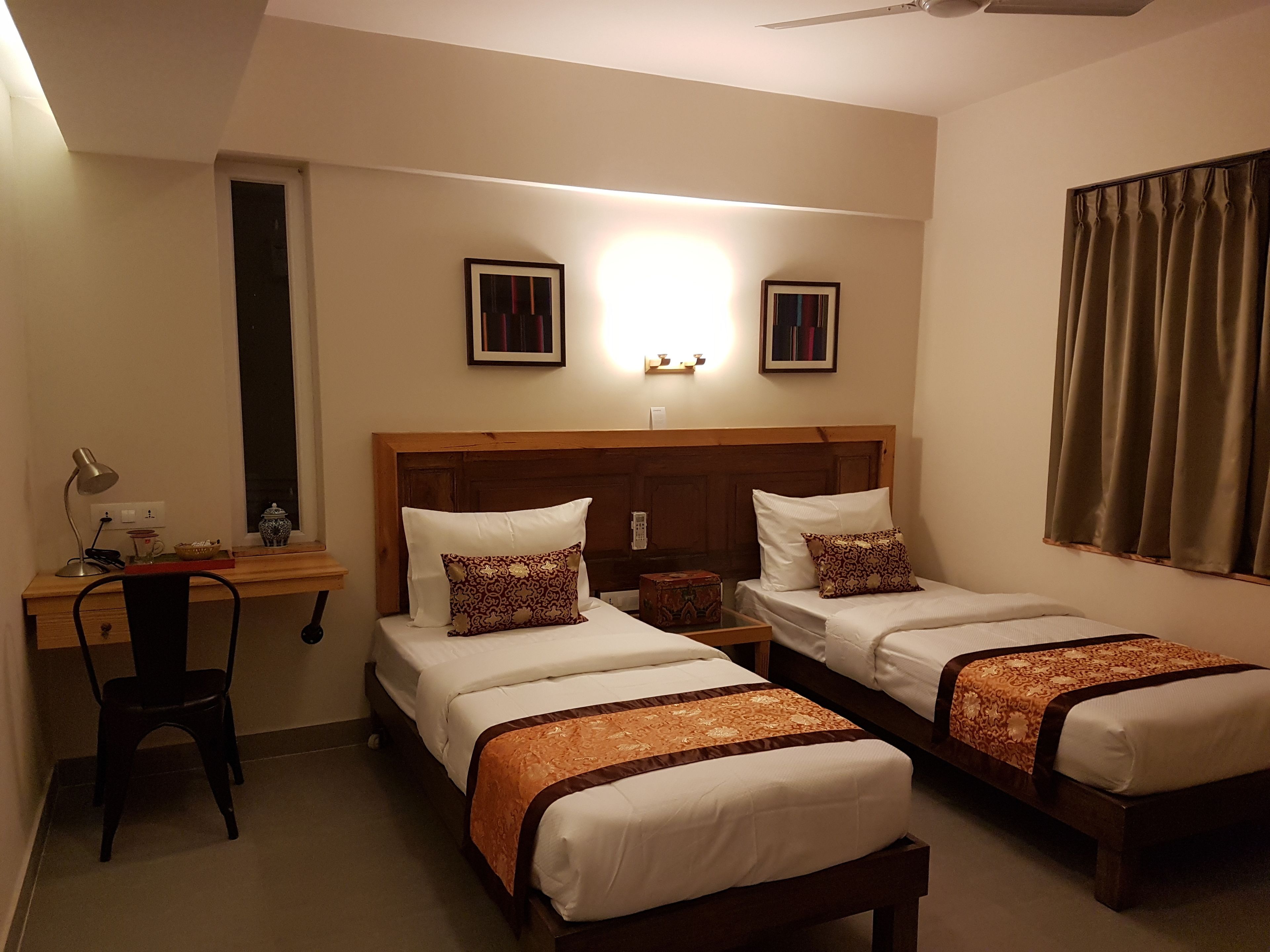 Une chambre d’hôtel avec deux lits simples, un bureau avec une chaise, une lampe et deux tableaux encadrés au mur.