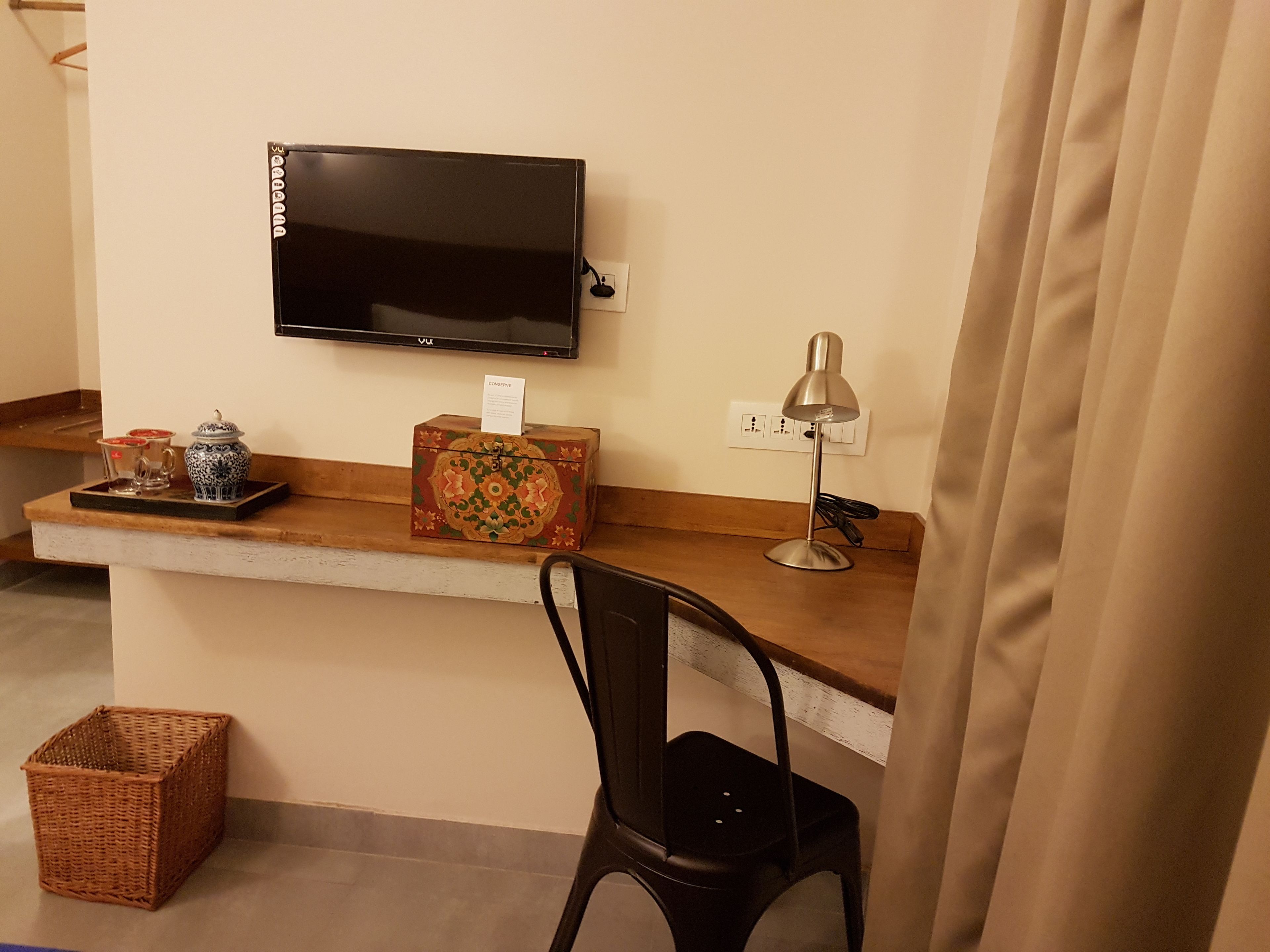 A small workspace with a TV mounted on the wall, a desk with a lamp, a chair, a decorative box, and a wicker basket.