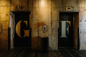 Zwei Aufzugtüren mit großen Buchstaben 'G' und 'F', eingelassen in eine abgenutzte Betonwand, mit einem kleinen Lautsprecher auf dem Boden.