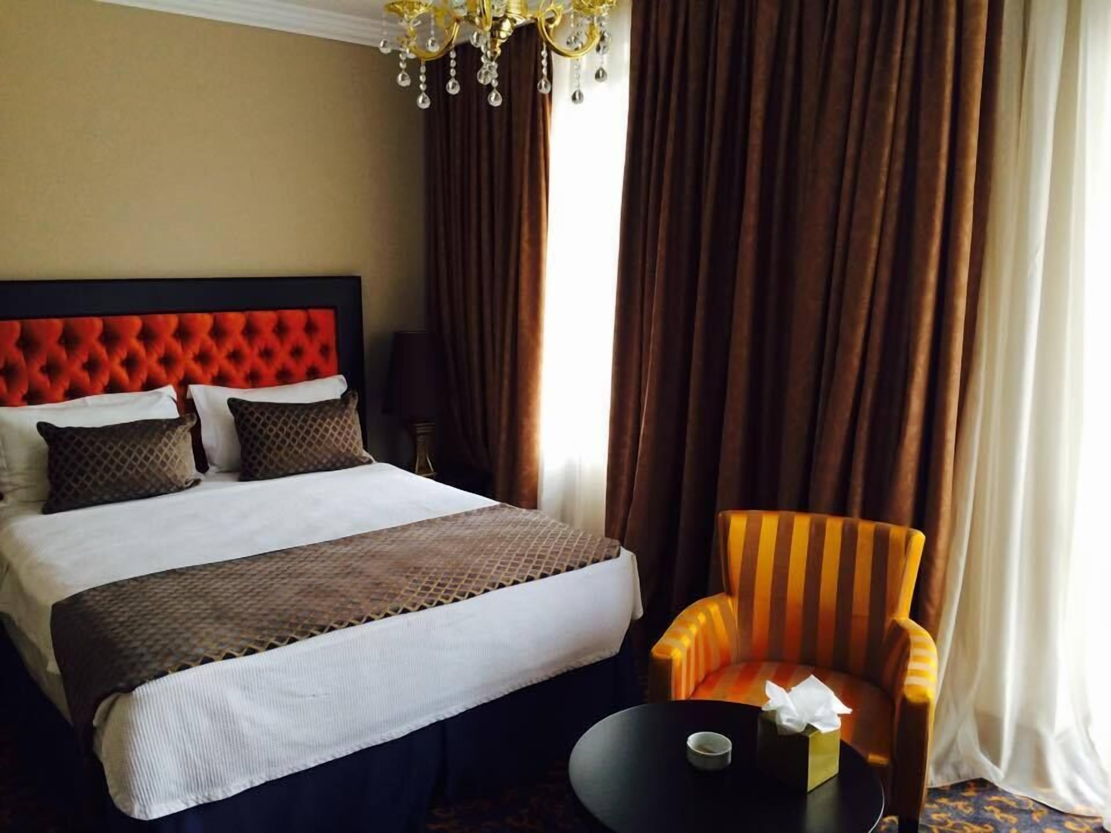 Una habitación de hotel con una cama grande, un cabecero capitonado, un sillón a rayas, una mesita con una taza y una araña.
