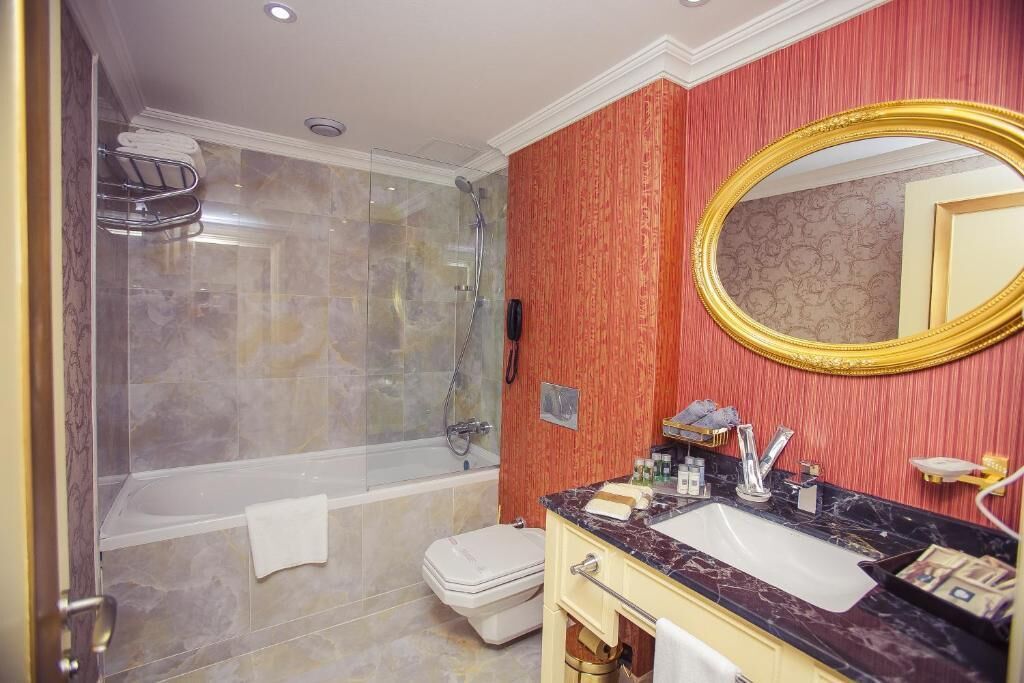 Une salle de bain dotée d’une douche à parois de verre, de toilettes blanches, d’un lavabo avec un plan de travail en marbre et d’un grand miroir encadré d’or.