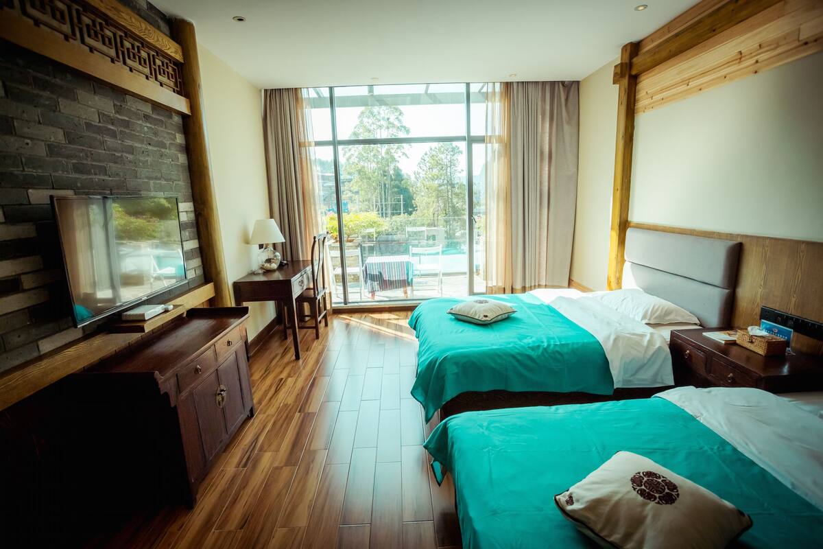 A hotel room with two beds, a TV, a wooden cabinet, and a large window offering a view of the outdoors.