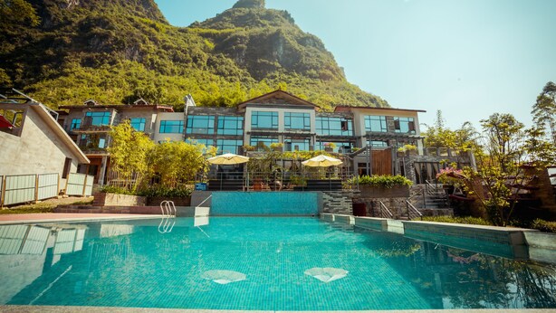 A hotel with a swimming pool, a mountain backdrop, and outdoor seating with umbrellas. - Moonlit Sky Resort (Guilin)