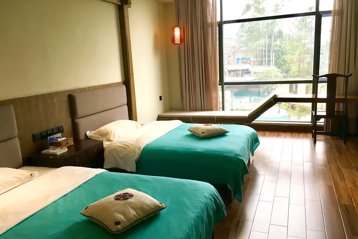 A hotel room with two beds, a window with a view of greenery, a wooden floor, and a nightstand with a lamp.
