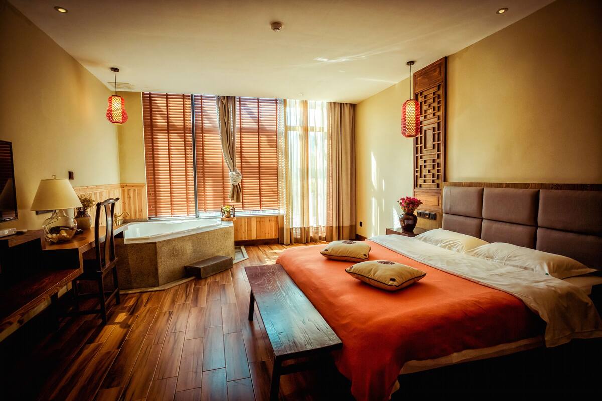 A hotel room with a large bed, a bench, a desk, a lamp, a window with blinds, and a wall-mounted shelf.