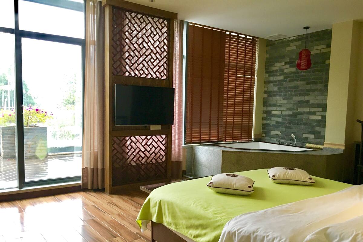 A hotel room with a large bed, a TV, a window with blinds, a bathroom with a bathtub, and a view of the outdoors.