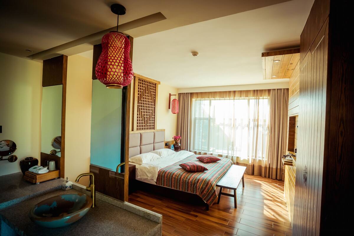 A bedroom with a large bed, a wooden floor, a bathroom with a sink, and a window with curtains.