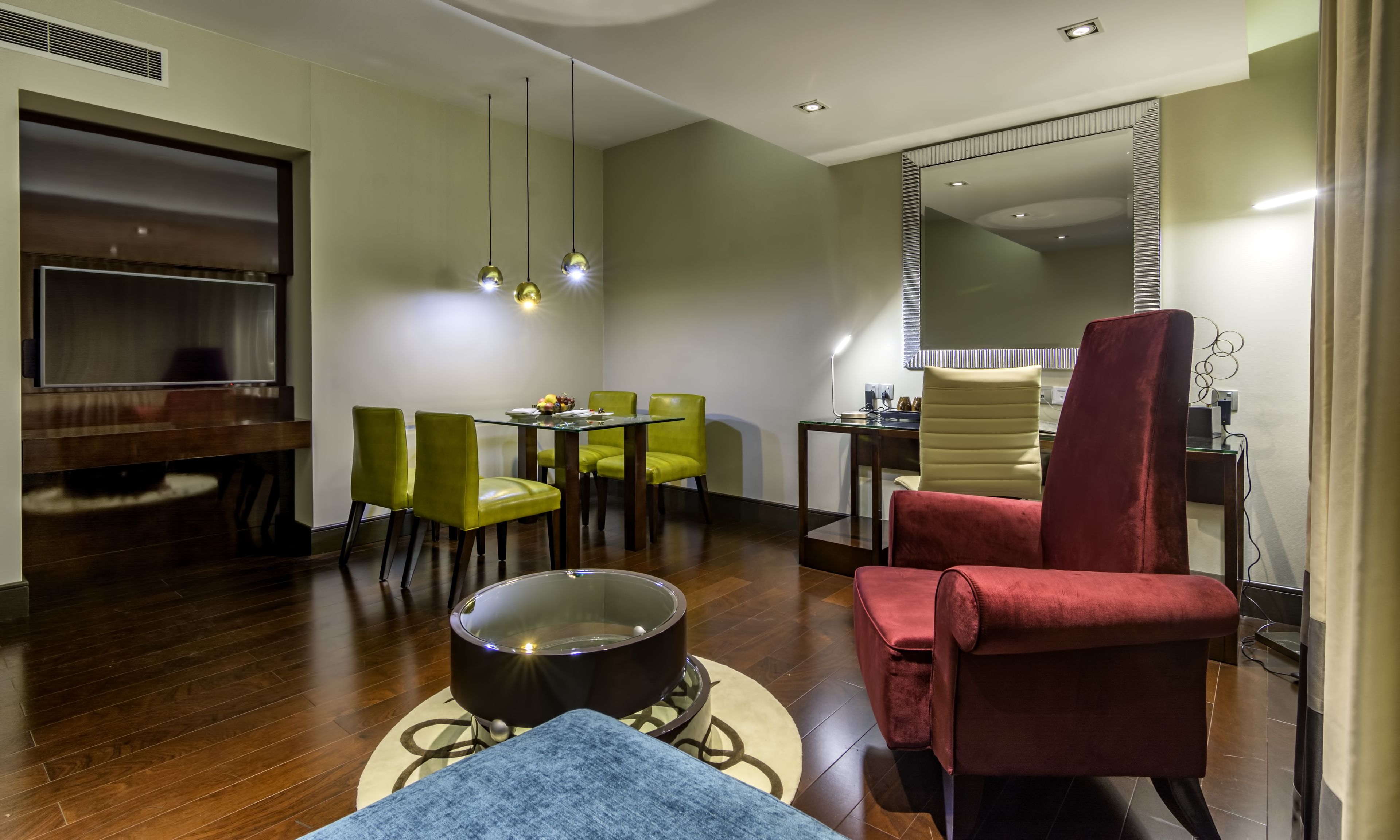 A modern living room with a flat-screen TV, green and red chairs, a round coffee table, and a large mirror.