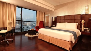 A modern hotel room with a large bed, a red chair, a round coffee table, and a view of the city through large windows. - The Raintree Dhaka (Dhaka)
