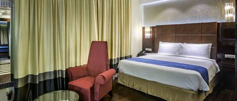 Habitación de hotel con una cama grande, un sillón rojo, una mesa de centro de vidrio y una cortina con diseño a rayas.