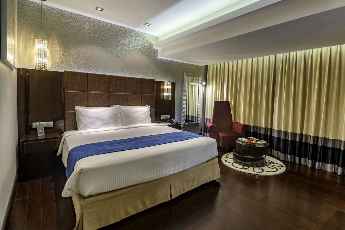 Ein Hotelzimmer mit einem großen Bett, einem Holzkopfteil, einem Nachttisch mit Lampe, einem Sessel und einem kleinen runden Tisch mit Früchten.