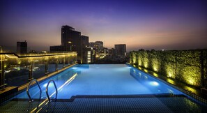 Piscina infinita en la azotea con vistas al horizonte urbano, zona iluminada junto a la piscina y barandilla de cristal.