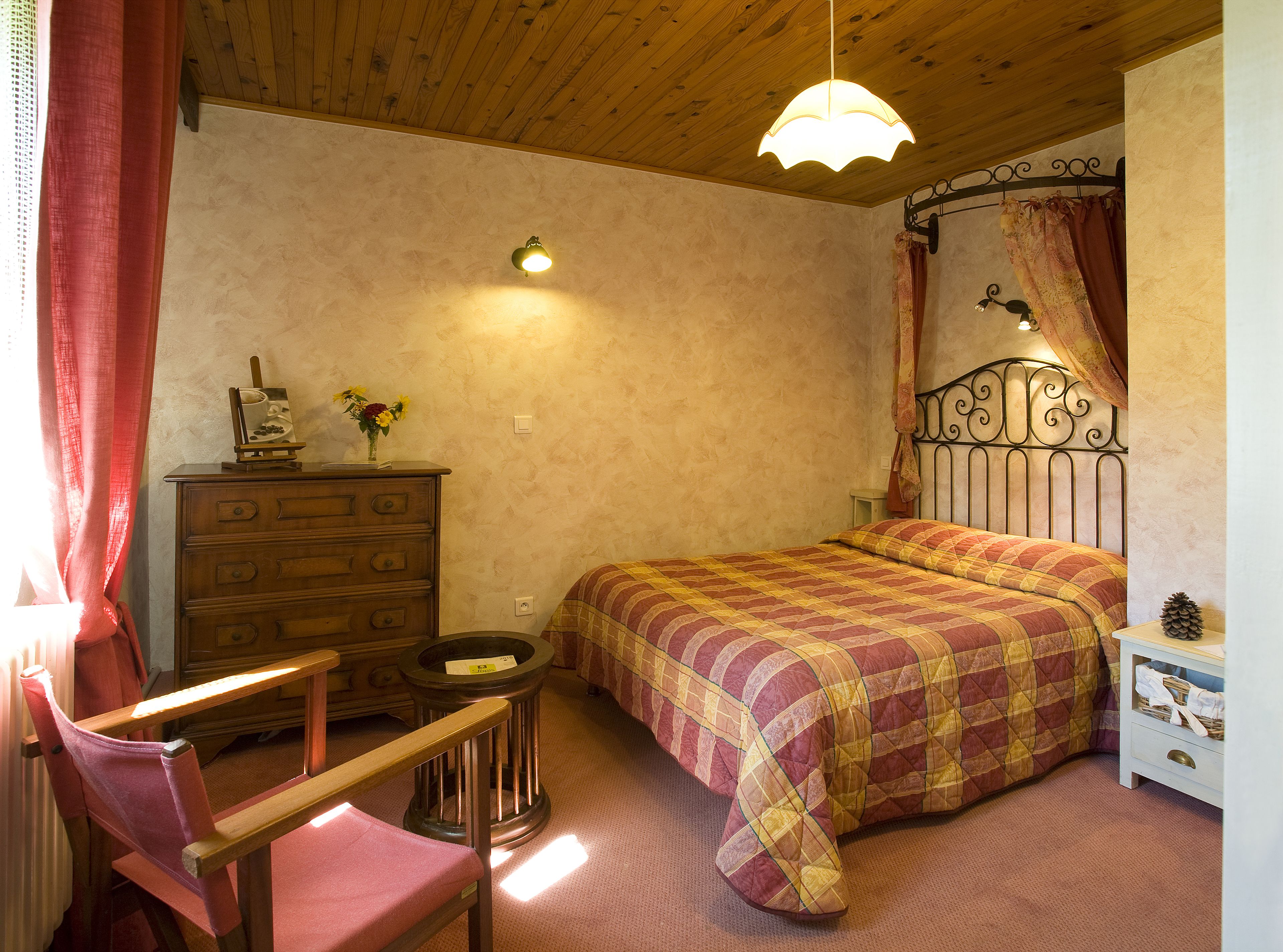 A bedroom with a wooden ceiling, a bed with a patterned bedspread, a dresser, a chair, and a small table with a lamp.