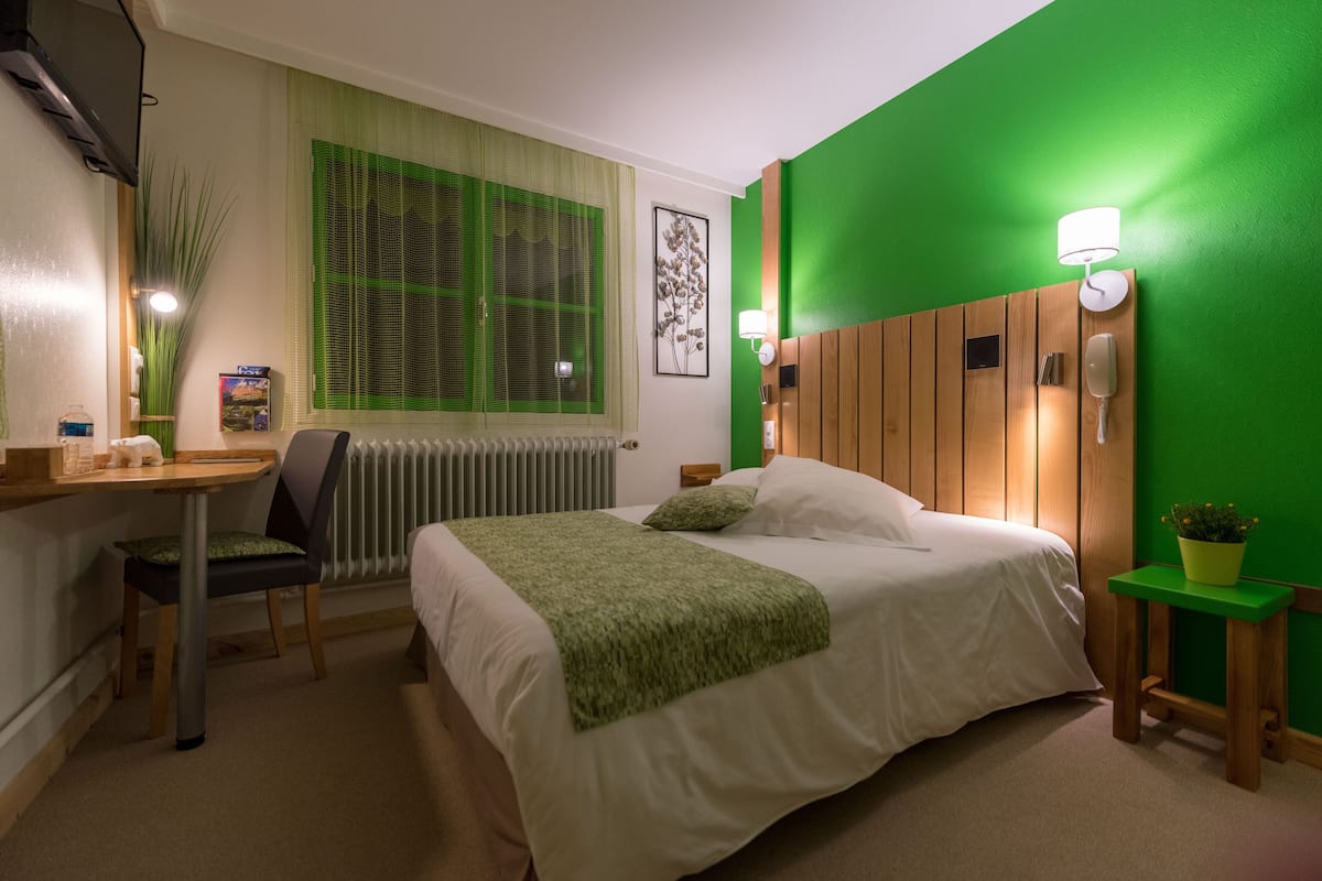 A hotel room with a bed, a desk, a chair, a radiator, a window with green blinds, a wall-mounted lamp, and a potted plant.
