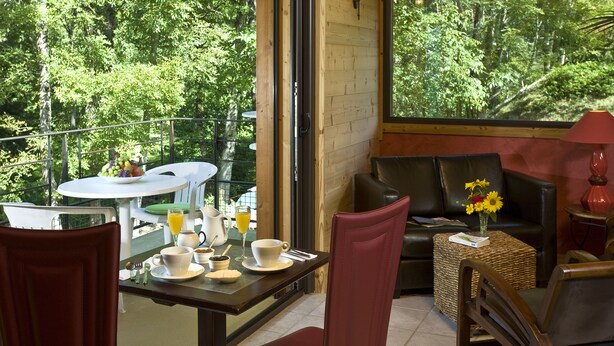 A dining area with a table set for breakfast, a sofa, and a view of a forest through large windows. - Hôtel Auberge du Morge (Les Echelles)