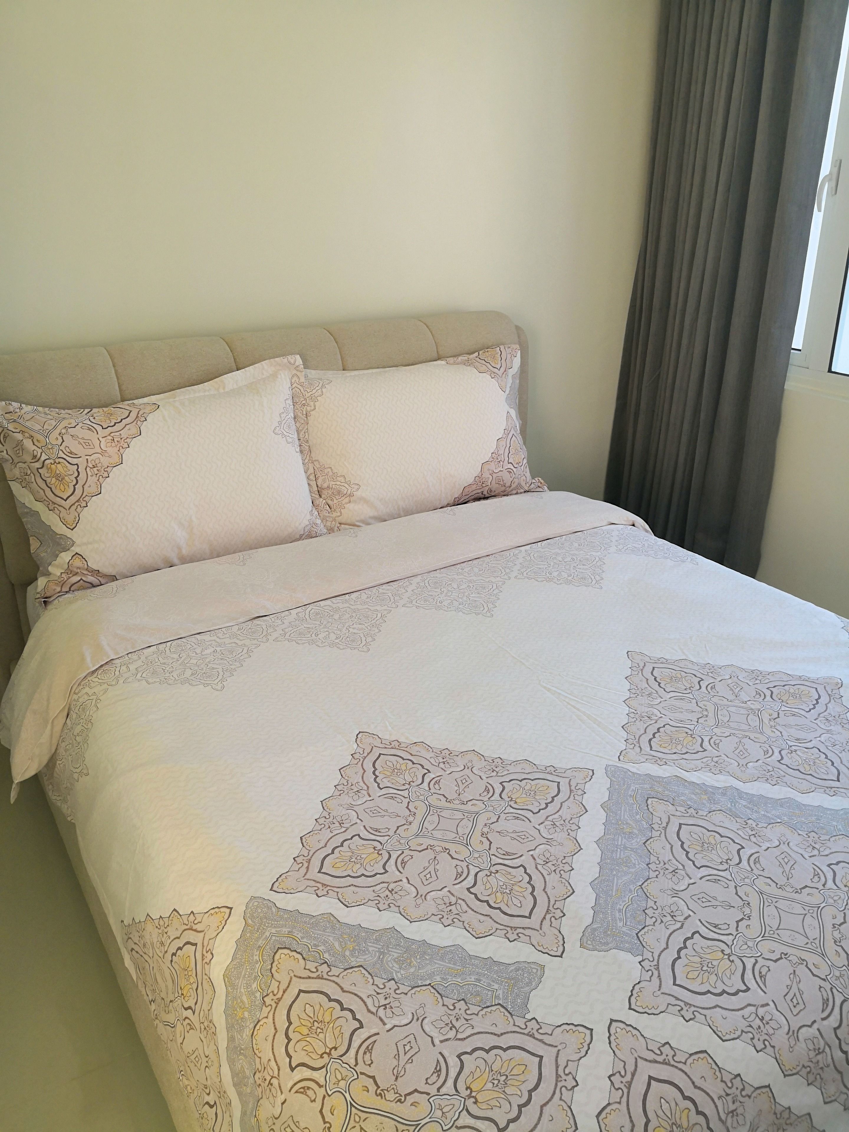 Ein Bett mit beigem Kopfteil, zwei Kissen mit Blumenmuster und einer Tagesdecke mit geometrischem Design.