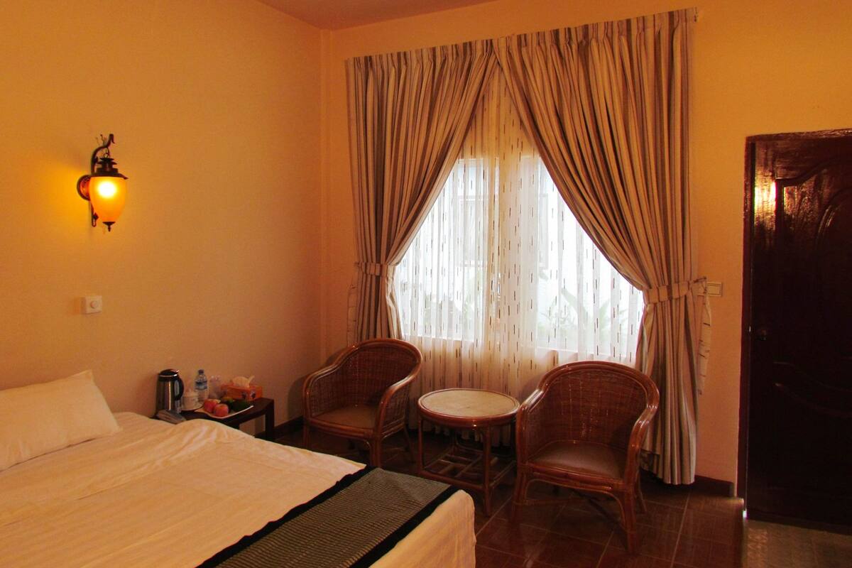 A hotel room with a bed, two wicker chairs, a small table, a wall-mounted lamp, and a window with curtains.