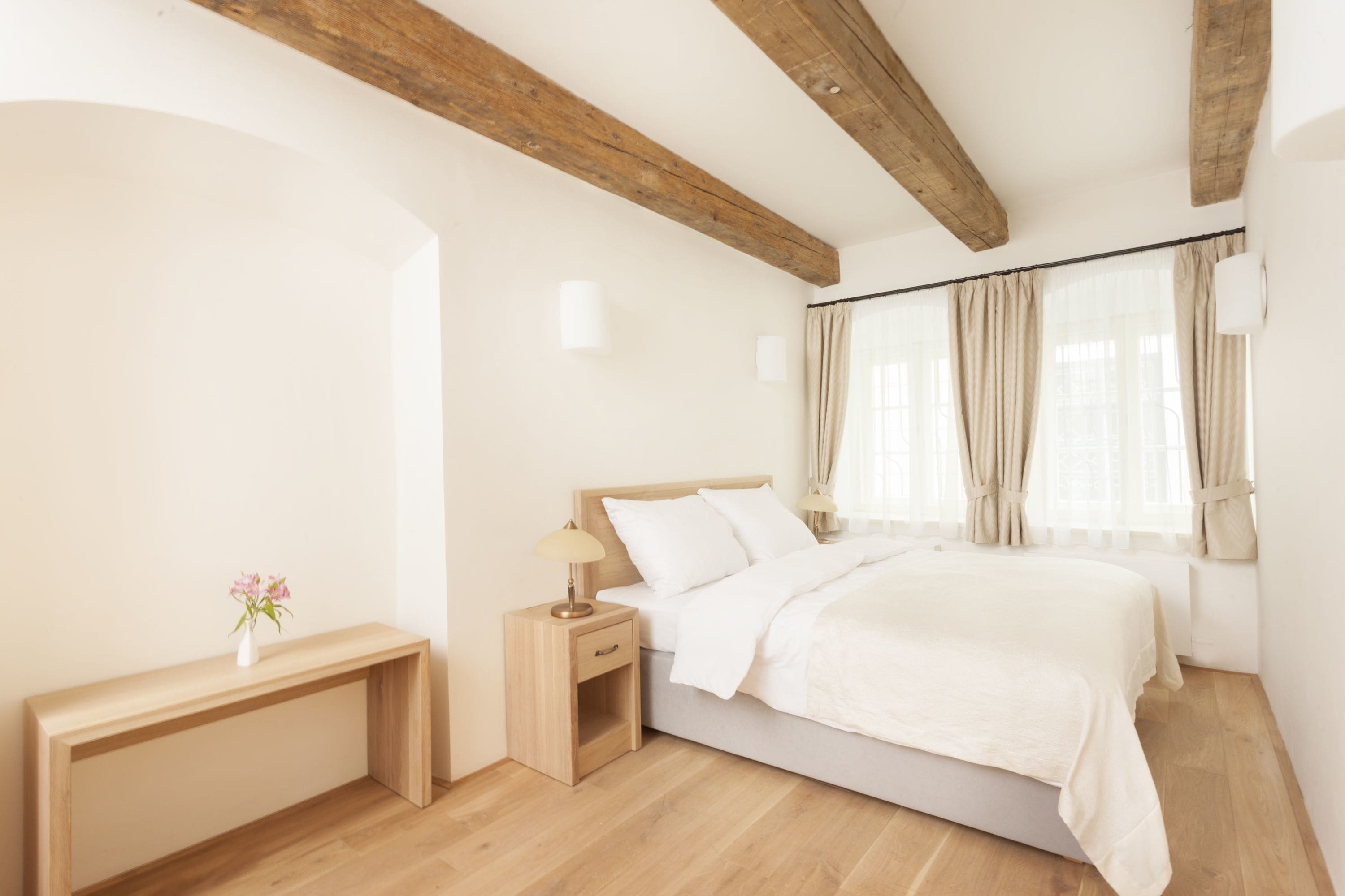 Een slaapkamer met een houten plafond, een bed met witte lakens, een nachtkastje met een lamp en een raam met gordijnen.
