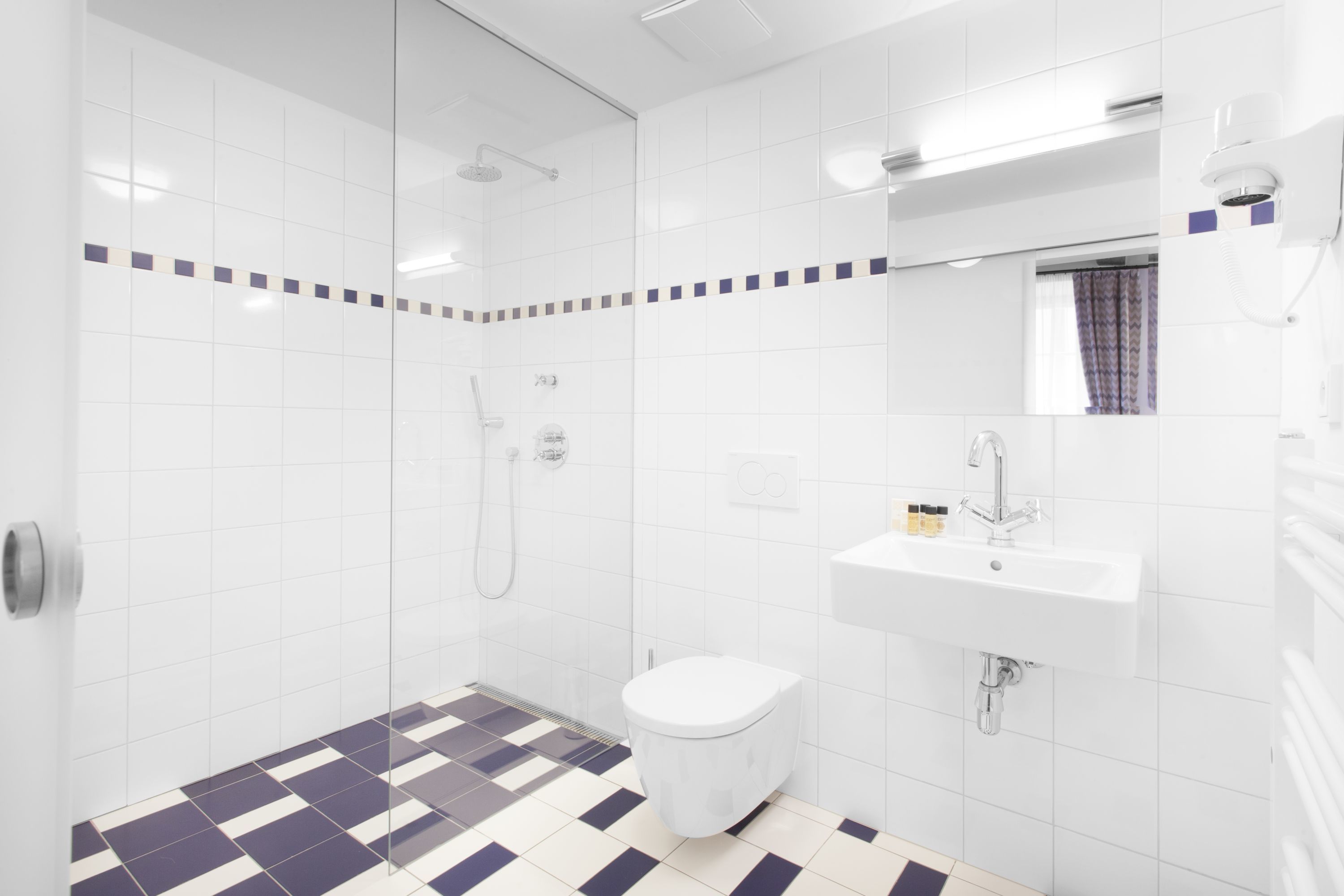 Un baño moderno con azulejos blancos, suelo de cuadros, inodoro blanco, lavabo y zona de ducha.