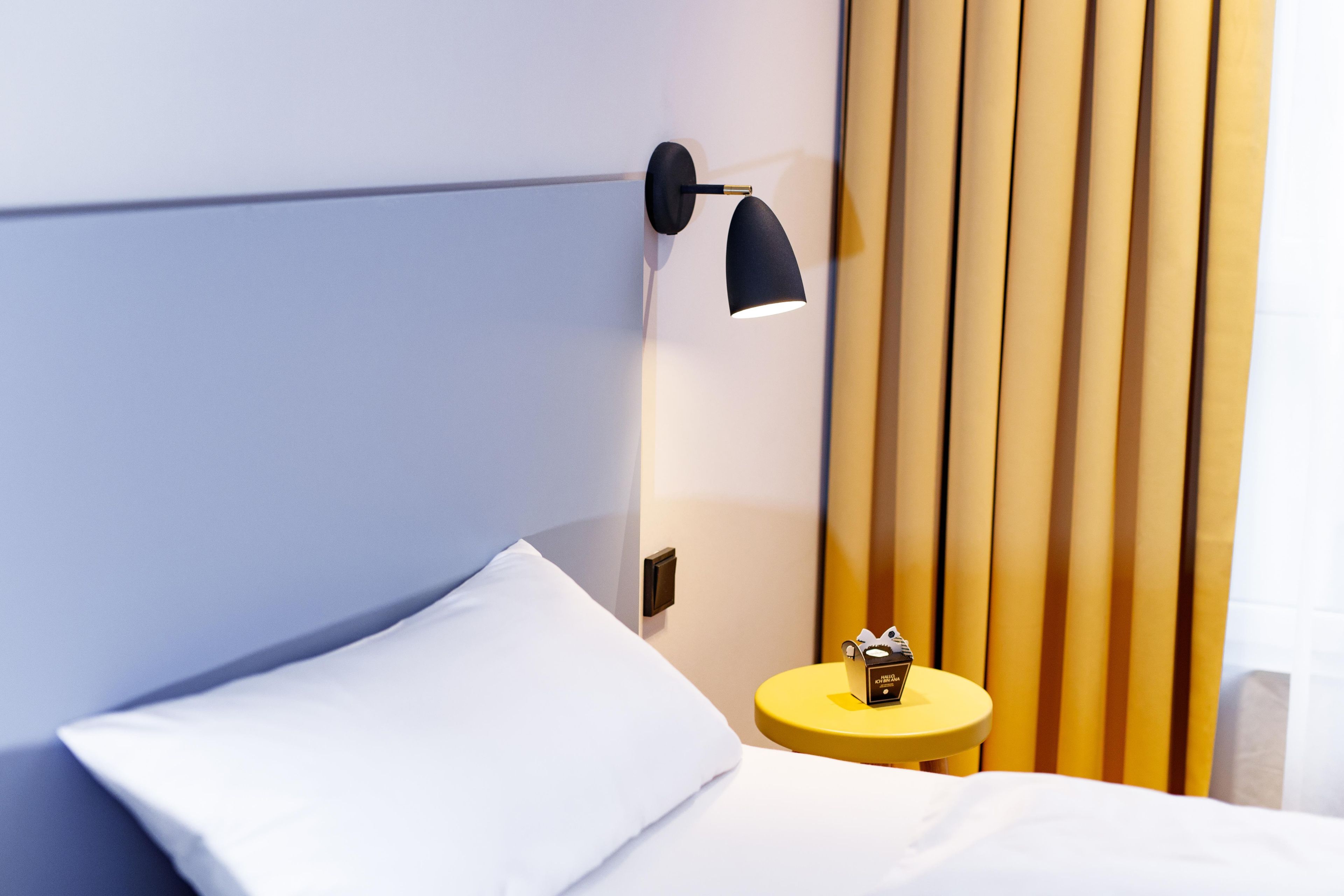A modern hotel room with a neatly made bed, a small round table with a lamp, and a wall-mounted light fixture.