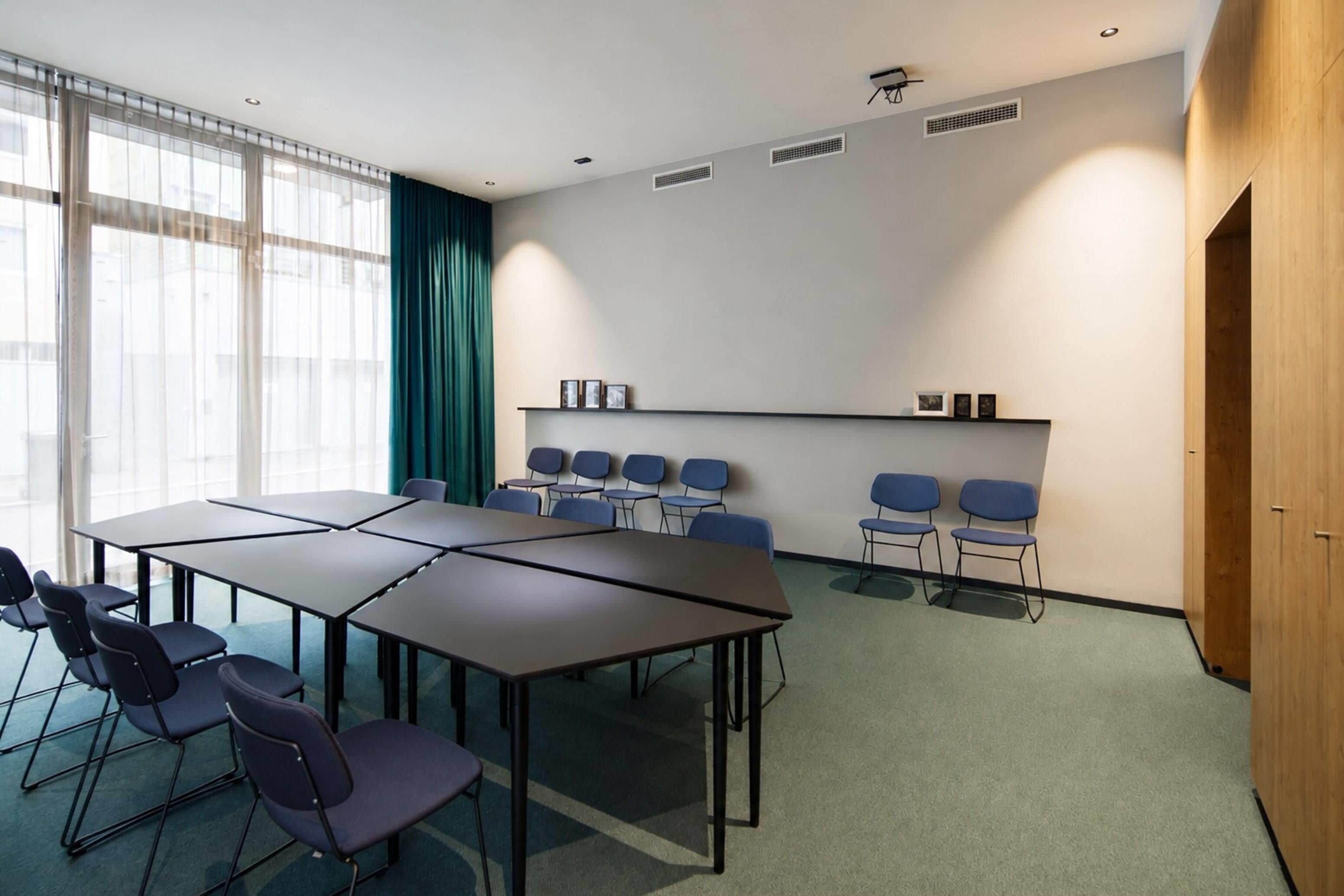 Un salón de conferencias con una gran mesa y sillas, una pantalla de proyector y un ventanal con cortinas.