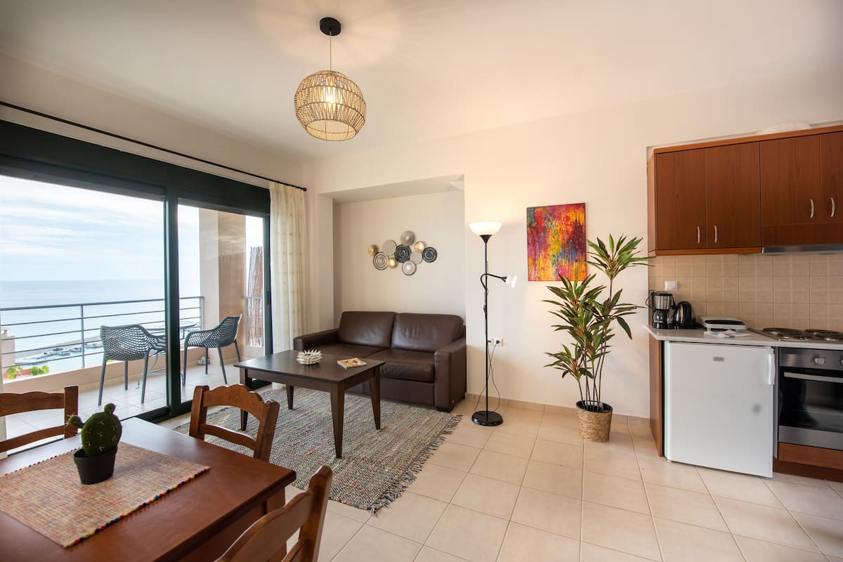 Un salon moderne avec un canapé, une table basse et des fauteuils, un balcon avec vue, une cuisine équipée de meubles et d’appareils électroménagers, et un lampadaire.