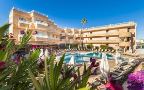 A hotel with a swimming pool surrounded by sun loungers and umbrellas, a palm tree, and a clear blue sky. - Mandisur Apartments (Manacor)