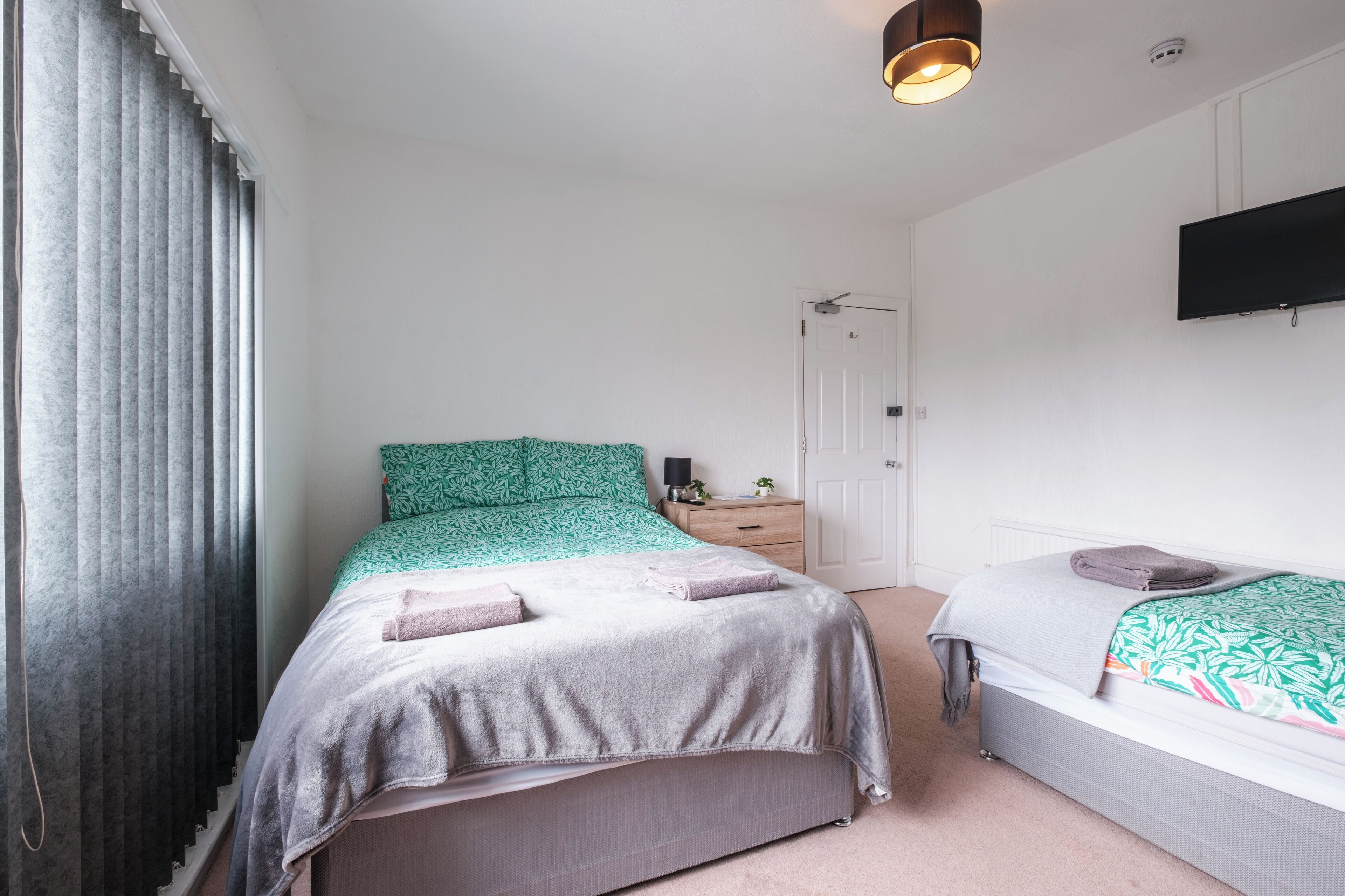 Un dormitorio con cama, mesita de noche, cómoda, televisor montado en la pared y una puerta.