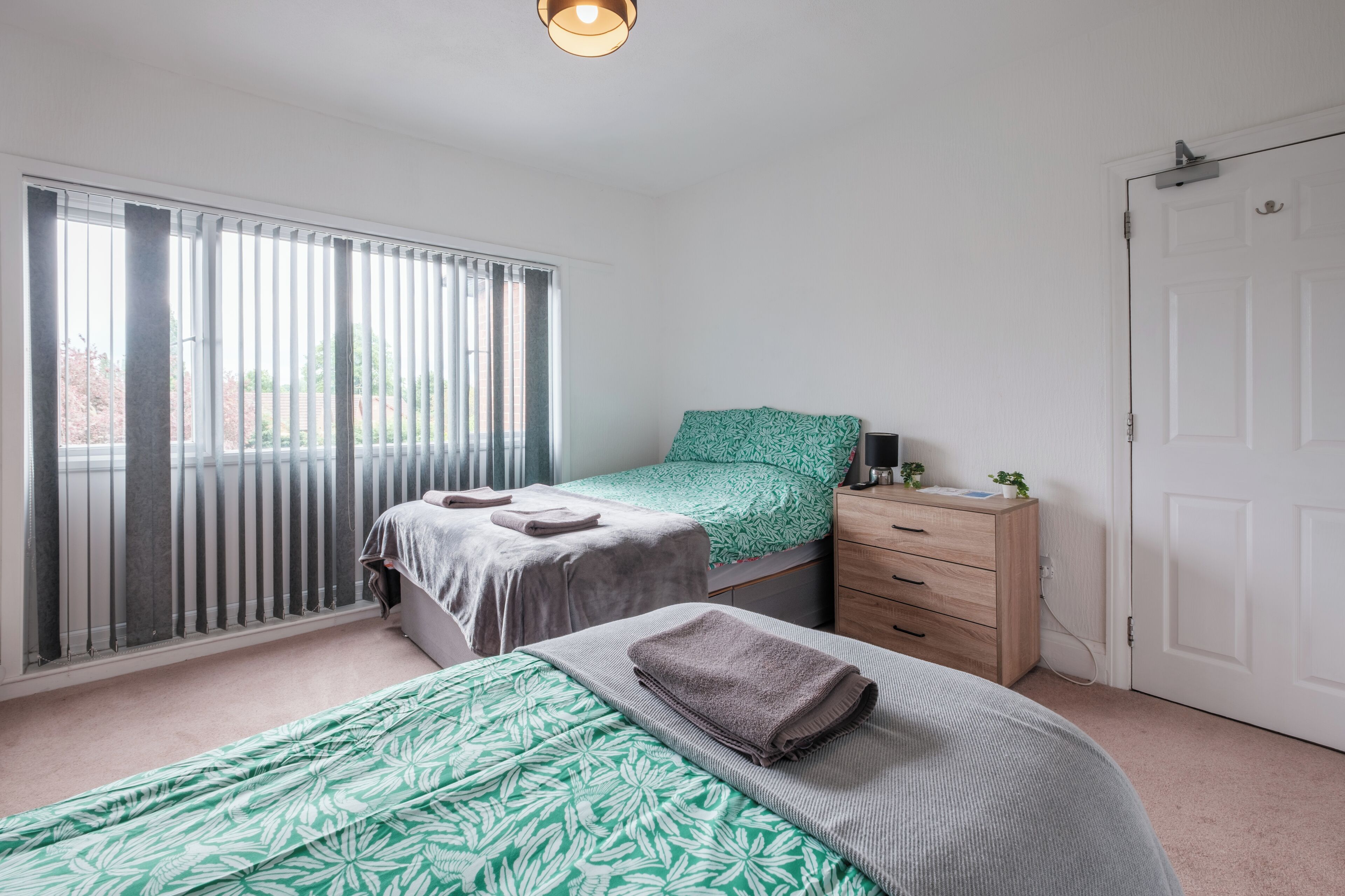 A bedroom with two beds, a window with blinds, a bedside table with a lamp, and a wardrobe.