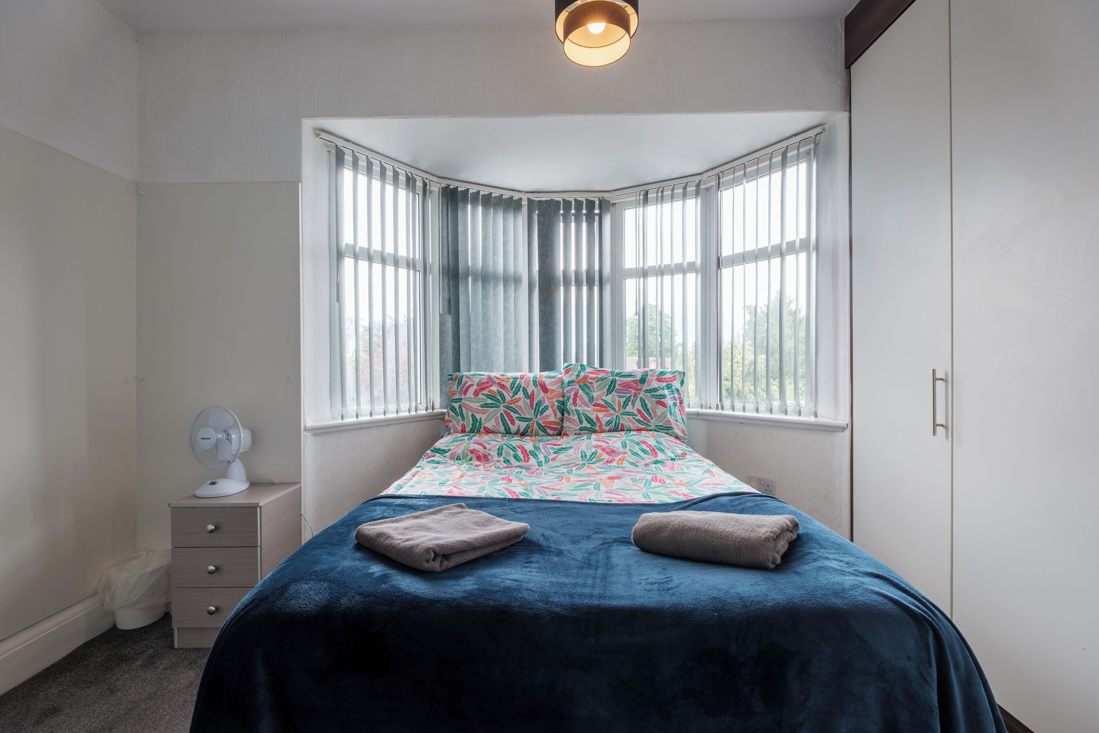 Un dormitorio con cama, mesita de noche, ventilador y ventana con cortinas.