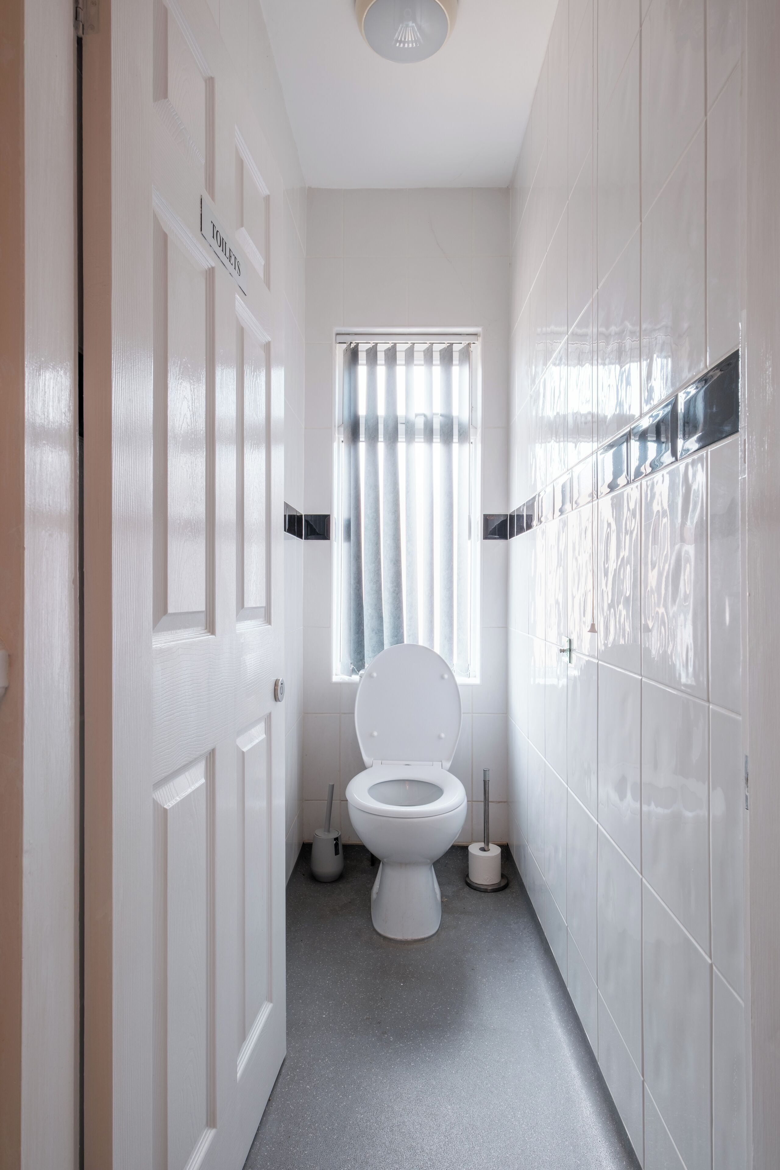 Un baño estrecho con inodoro, una ventana con cortinas y una puerta con la leyenda 'Toilets'.