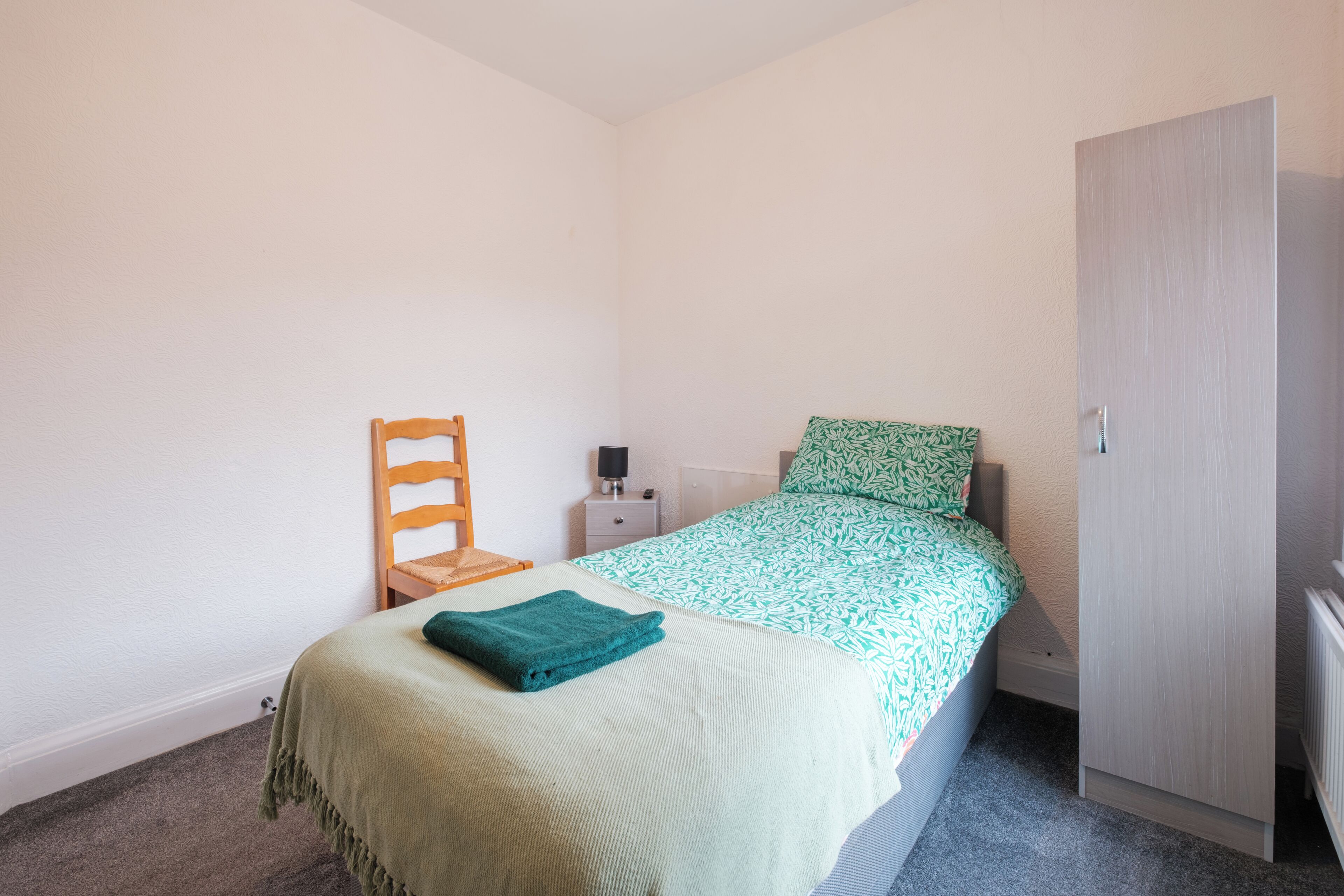 Un dormitorio pequeño y bien iluminado con una cama individual, una silla de madera, una mesita de noche y un armario.