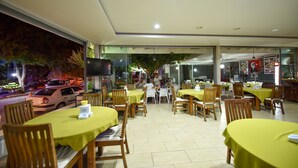 A restaurant with tables covered in yellow tablecloths, wooden chairs, and a view of a street with parked cars and trees outside. - Lidya City Hotel (Fethiye)
