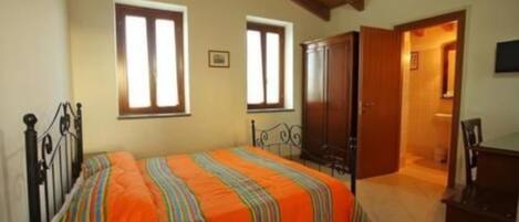 Un dormitorio con cama, dos ventanas, un armario y una puerta que da a otra habitación.