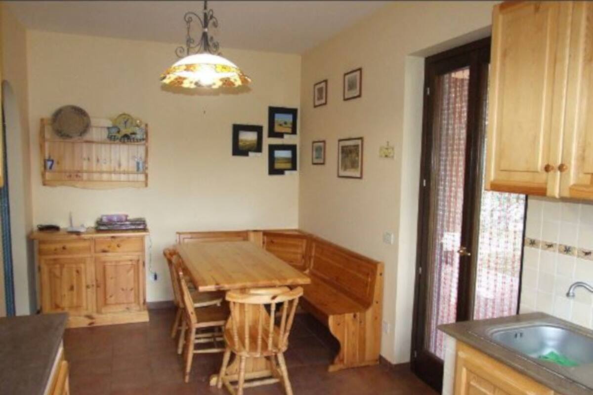 Een kleine keuken met houten kastjes, een eettafel met stoelen en een hangende lamp.