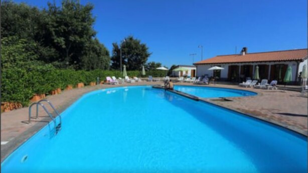 Une piscine entourée de verdure, agrémentée de chaises longues et de parasols, et bordée d’un bâtiment dont le toit est recouvert de tuiles.