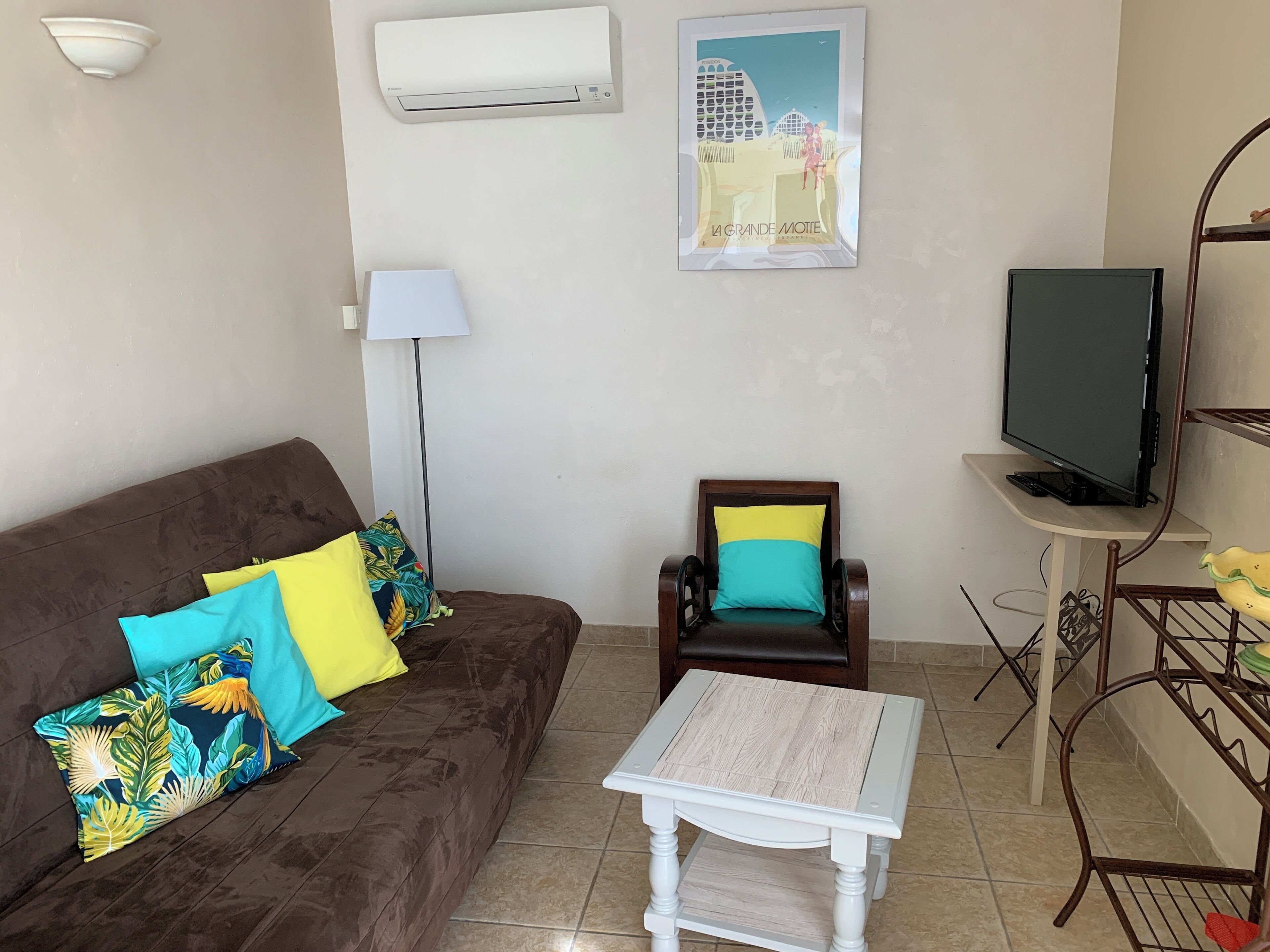 A living room with a brown sofa, a wooden chair, a small white table, a TV on a stand, and a poster on the wall.