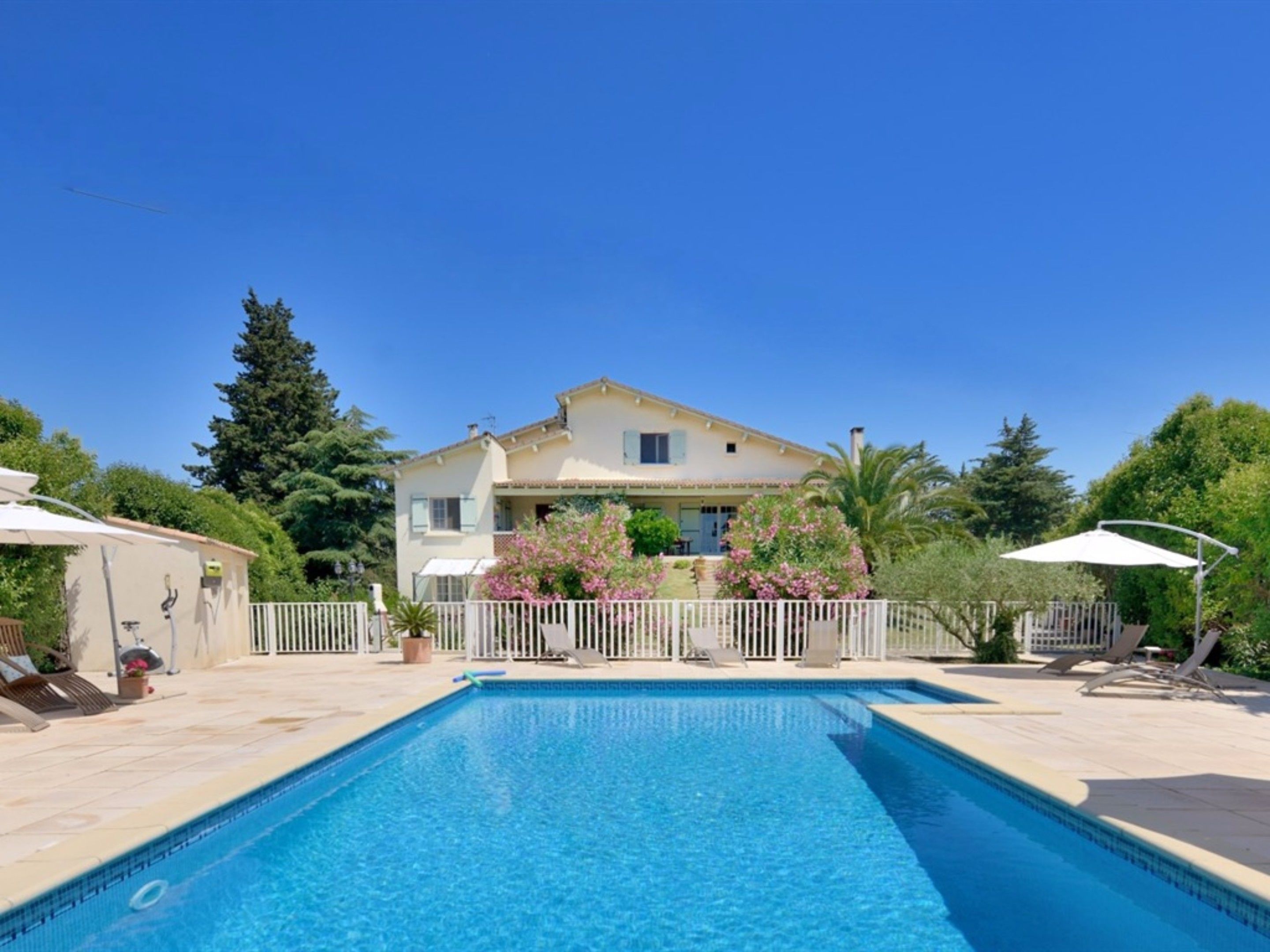Una piscina con una valla blanca, una casa al fondo y follaje circundante.