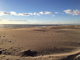 Una extensa playa arenosa con huellas y rocas, cielo despejado y una vista distante del océano.