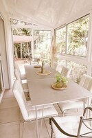 Un invernadero con una larga mesa de comedor, sillas blancas y un jarrón con plantas.