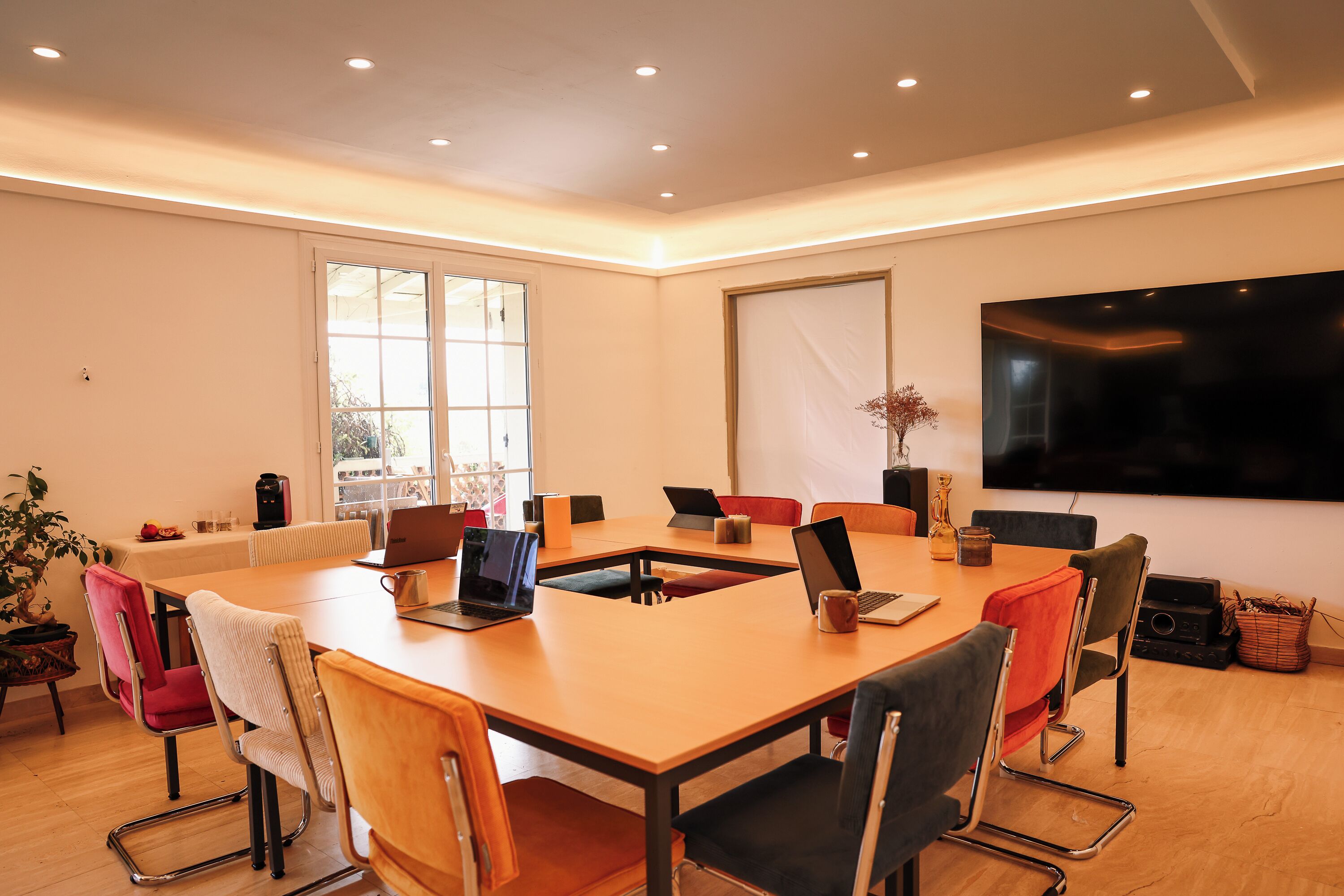 A modern meeting room with a large table, multiple chairs, laptops, and a flat-screen TV.