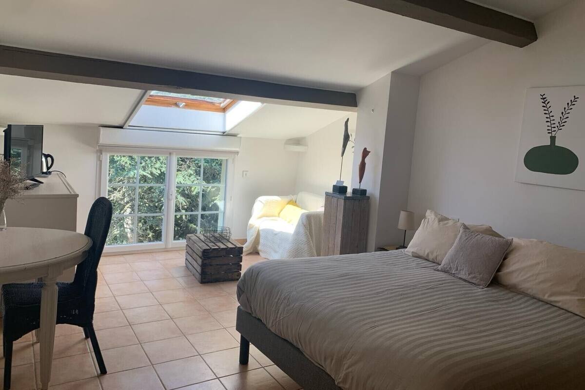 A bedroom with a bed, a desk, and a chair. There is a skylight above the bed and a window in the background.