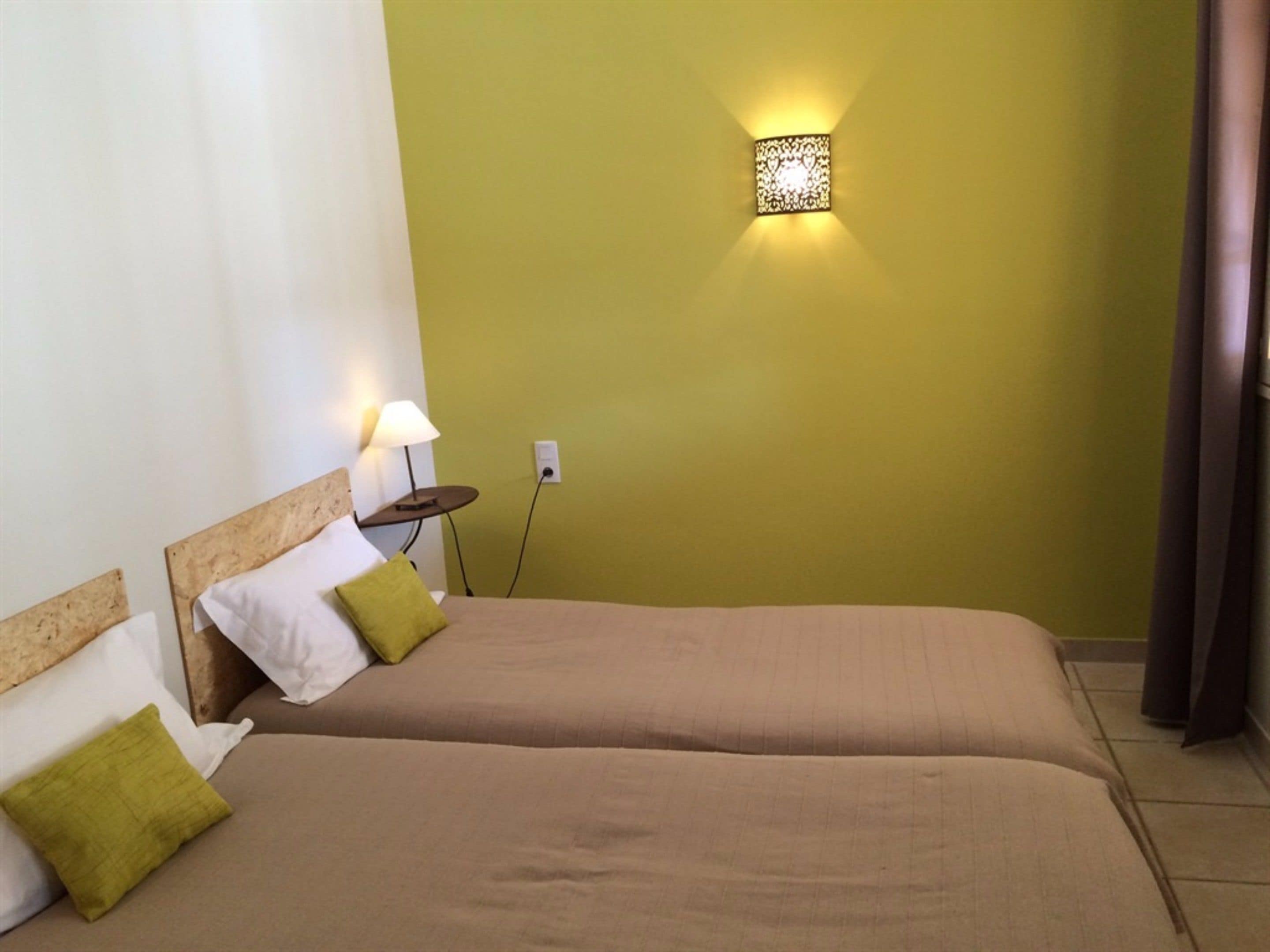 A hotel room with two beds, a nightstand with a lamp, and a wall-mounted light fixture.