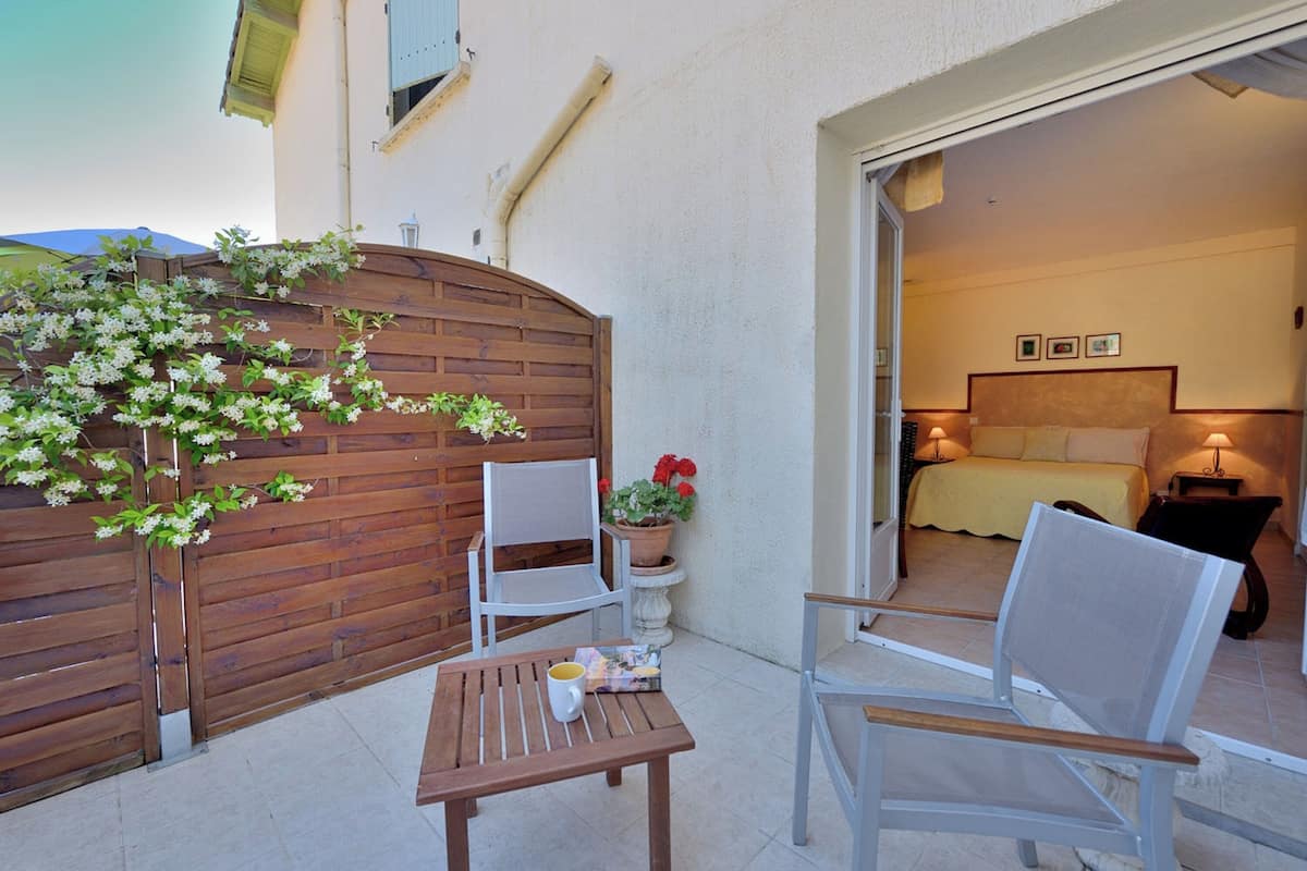 A patio with a wooden table and chairs, a potted plant, and a view into a bedroom with a bed and bedside tables.
