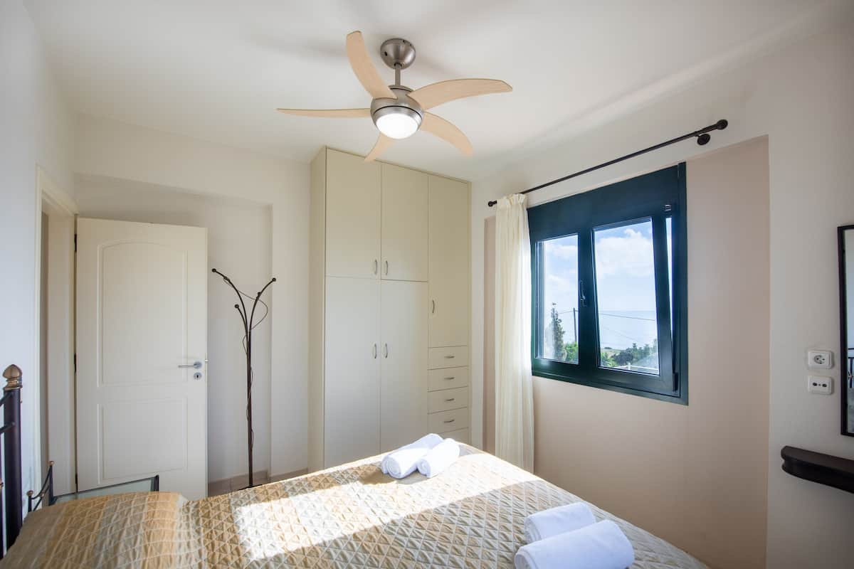 A bedroom with a bed, a ceiling fan, a wardrobe, a window with a view, and a door.