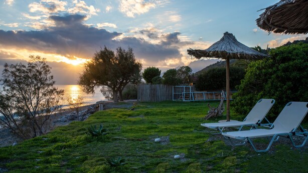 Zona exterior con césped, sillas de descanso y un paraguas de paja, con vista a un cuerpo de agua y un atardecer de fondo.