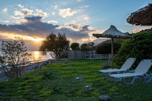 Zona exterior con césped, sillas de descanso y un paraguas de paja, con vista a un cuerpo de agua y un atardecer de fondo.