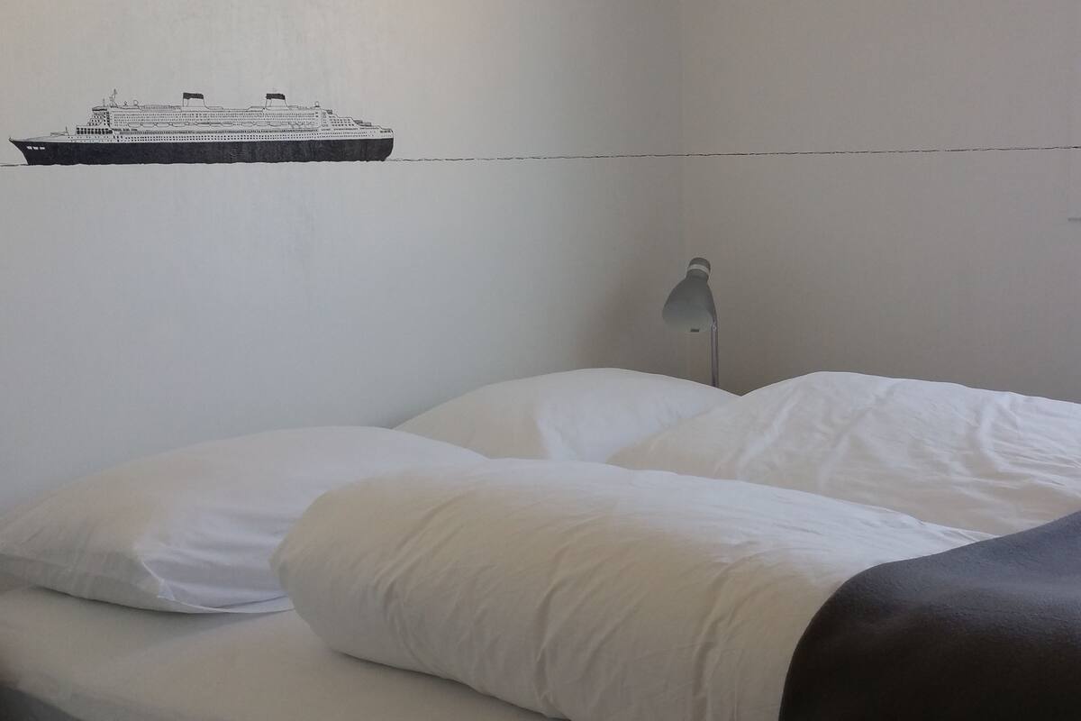 Una cama con ropa de cama blanca y una manta oscura, una lámpara y un adhesivo mural de un barco.