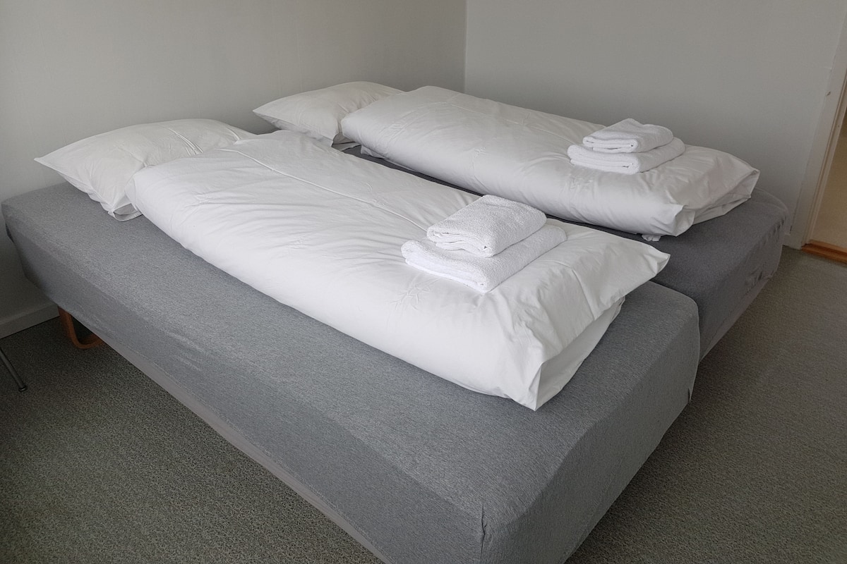 Una habitación con dos camas individuales, cada una con sábanas blancas y almohadas, y un protector de colchón gris.