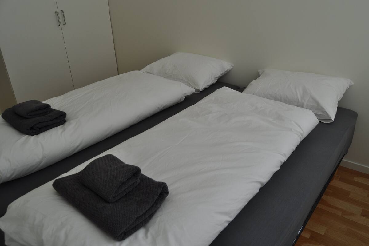 Habitación doble con dos camas individuales, cada una con ropa de cama blanca y toallas grises oscuras.